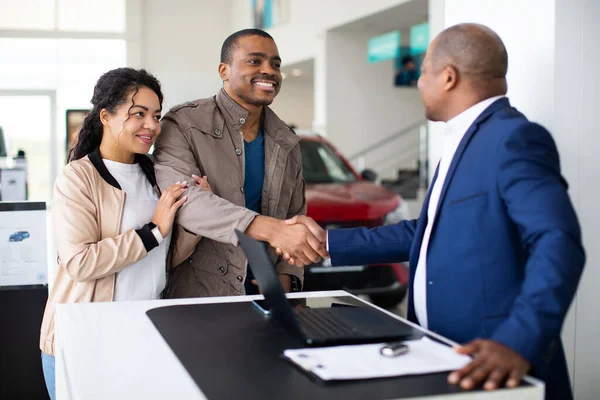 Smiling African American couple shaking hands with car dealer at desk, celebrating successful purchase agreement, lifestyle moment of auto shopping, finance and trust