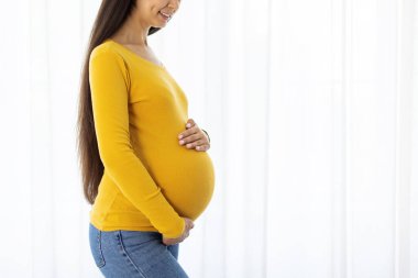 Side view of young pregnant woman in yellow top at home, smiling and holding belly with care. Bright prenatal lifestyle concept, simple and modern, copy space