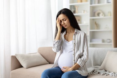 Pregnant woman at home touching head and holding belly, looking unwell. Headache or discomfort during pregnancy, authentic maternity lifestyle moment indoors