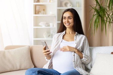 Smiling pregnant woman sitting on sofa using smartphone, hand on belly. Bright prenatal lifestyle image showing technology, modern comfort and anticipation