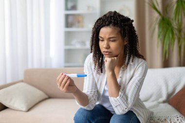 Endişeli Afro-Amerikan kadın evinde koltukta oturuyor, hamilelik testi yapıyor ve ciddi bir ifadeyle sonuçlara bakıyor, aile planlaması konusunda kararsız.
