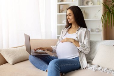 Smiling pregnant woman using laptop at home while holding belly. Casual maternity lifestyle portrait showing modern balance of work and motherhood