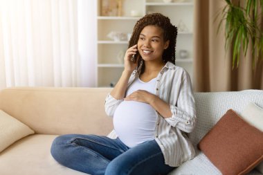 Gülümseyen hamile Afrikalı Amerikalı kadın koltukta oturuyor, telefonla konuşuyor ve karnını tutuyor, doğum sırasında yaşam tarzı bağlantısından ve aile beklentilerinden zevk alıyor.