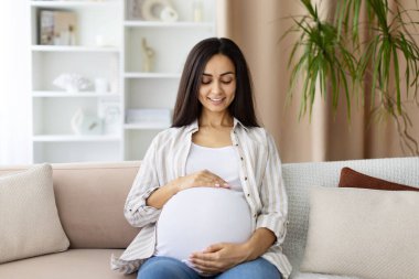 Smiling pregnant woman sitting on sofa, looking down at her belly with joy. Peaceful maternity lifestyle scene at home, celebrating motherhood and prenatal care