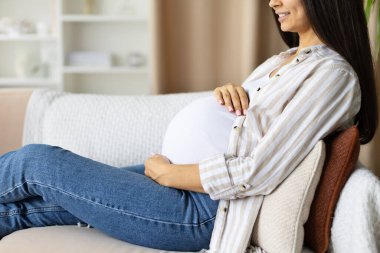 Closeup of pregnant woman relaxing on sofa at home, hand gently touching belly. Peaceful prenatal lifestyle and maternity moment, calm anticipation of motherhood, side view