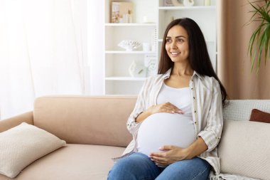 Pregnant woman at home sitting on sofa, smiling and holding belly with both hands. Bright prenatal lifestyle portrait with joyful anticipation of motherhood