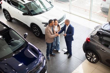 Happy African American couple shaking hands with car dealer in modern showroom, celebrating successful purchase, lifestyle and finance moment of auto shopping