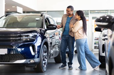 Smiling African American couple looking at car in showroom, lifestyle scene of shopping for new SUV, transport choice and family decision about modern vehicle purchase