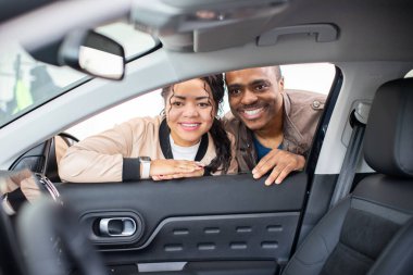 Smiling African American couple peeking inside car window in showroom, lifestyle moment of excitement, love and shopping for new vehicle, transport and ownership