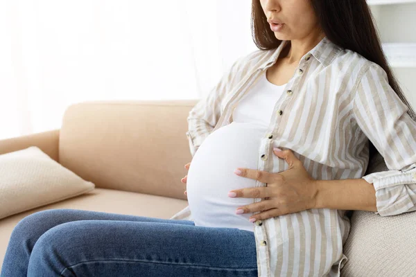 Closeup of pregnant woman sitting on couch at home, holding belly with both hands and breathing deeply. Real prenatal moment of motherhood and maternity care
