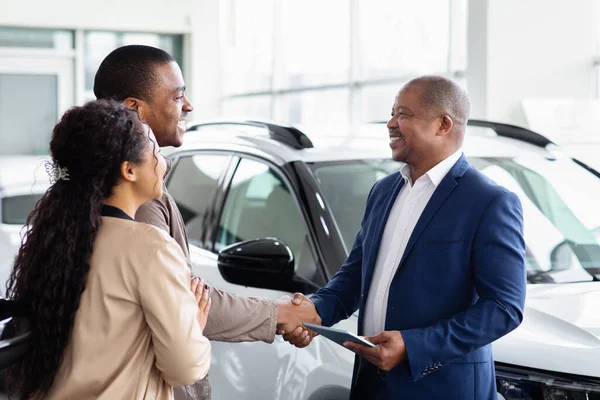 Smiling black couple handshaking with car dealer in showroom, symbol of trust, successful sale, lifestyle moment of new vehicle purchase and ownership