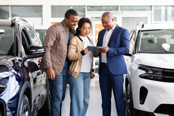 Car dealer showing digital tablet to smiling African American couple in showroom, reviewing new SUV details, lifestyle and business concept of purchase and transport