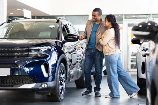 Smiling African American couple looking at car in showroom, lifestyle scene of shopping for new SUV, transport choice and family decision about modern vehicle purchase