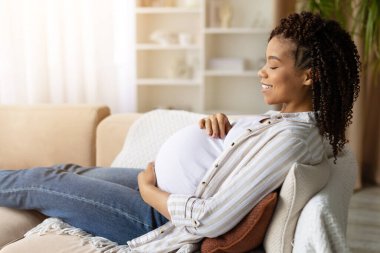 Side view of relaxed African American pregnant woman relaxing on cozy couch with eyes closed, smiling and touching belly. Peaceful prenatal rest and gentle home lifestyle with soft daylight