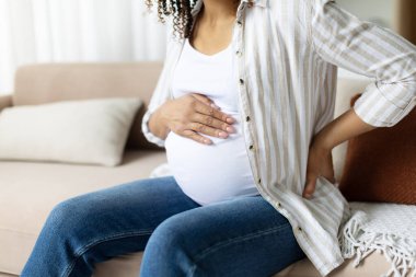 Closeup of pregnant woman holding hands on belly and touching back while sitting. Symbol of discomfort, prenatal body strain, and maternity challenges