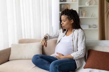 Pregnant African American woman checks smartwatch while seated cross-legged on sofa, one hand on belly. Timing contractions, prenatal monitoring, and planning in a cozy living room at home