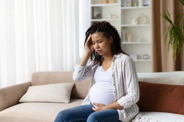 Thoughtful pregnant woman sitting on sofa, touching her head with concern. Concept of stress, prenatal headache, and maternity health, copy space