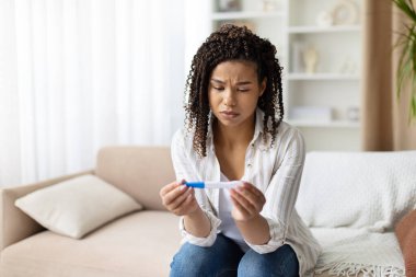Thoughtful African American woman looks at pregnancy test at home, feeling uncertain and concerned. Concept of decision making, fertility, and prenatal awareness