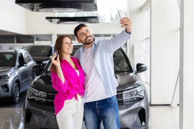 Smiling couple taking selfie with new car keys at dealership, man and woman celebrate vehicle purchase inside showroom. Lifestyle, technology and transport