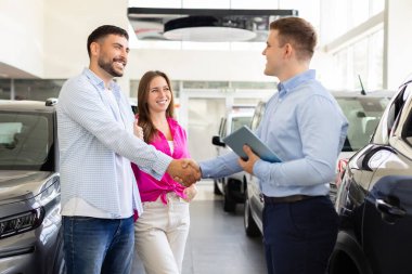 Happy couple shaking hands with car dealer inside modern showroom. Smiling man and woman making vehicle purchase decision at dealership center, closeup