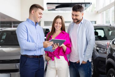 Salesman showing digital tablet to smiling couple at car dealership, man and woman review vehicle purchase details inside modern showroom. Lifestyle and transportation