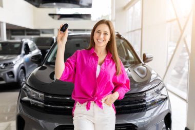 Smiling young woman standing in front of new car holding keys at dealership, confident customer celebrating purchase. Lifestyle, retail and transportation