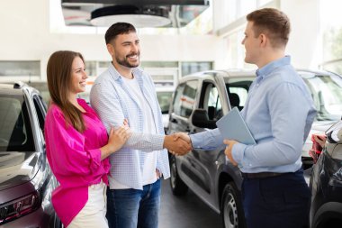 Smiling couple shaking hands with car dealer at showroom. Man and woman making agreement for new vehicle purchase, trust, lifestyle and transportation concept