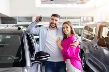 Happy couple posing with car keys near new vehicle at dealership, smiling man and woman hugging and celebrating purchase. Lifestyle, transport and ownership
