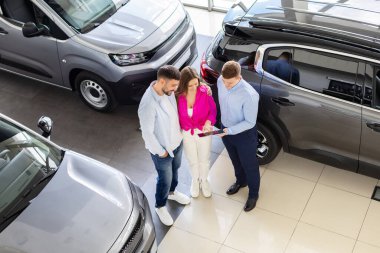 Car dealer showing details on tablet to smiling couple at dealership, above view. Man and woman standing between cars reviewing purchase agreement