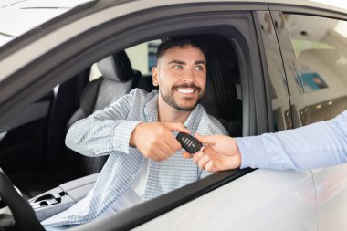 Smiling man in drivers seat of new car receiving keys at dealership, happy customer celebrating purchase and ownership. Lifestyle, transport and trust