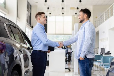 Smiling car dealer shaking hands with customer inside showroom, young man finalizing automobile purchase agreement. Lifestyle, trust, transport and consumer finance