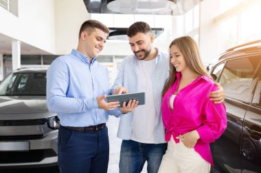Smiling car dealer showing tablet to happy couple at dealership. center, man and woman review automobile purchase details inside showroom. Lifestyle, consumer trust and transport