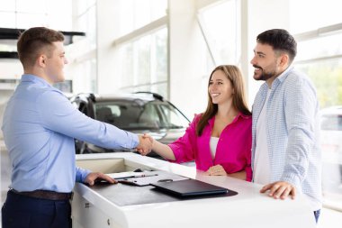 Happy couple shaking hands with car dealer at dealership desk after signing papers, smiling man and woman finalize automobile purchase. Trust, lifestyle and transport
