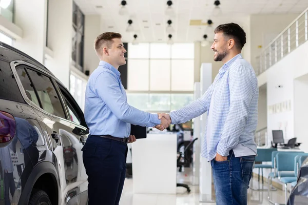 Smiling car dealer shaking hands with customer inside showroom, young man finalizing automobile purchase agreement. Lifestyle, trust, transport and consumer finance