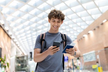 Young Caucasian man with backpack holding passport and smartphone, smiling inside airport terminal. Travel concept of departure, boarding, youth lifestyle and digital routine