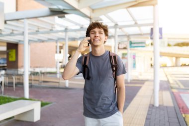 Young Caucasian man walking at airport while talking on phone and smiling. Represents casual travel lifestyle, communication, and happy youth on the go