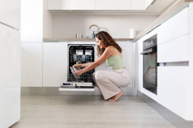Young Caucasian woman loading dishwasher at home kitchen. Concept of domestic chores, modern lifestyle, home appliance use, and everyday household routine