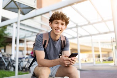 Young Caucasian man with backpack smiling at camera while holding smartphone at airport. Lifestyle image of youth, modern travel, and positive journey vibe