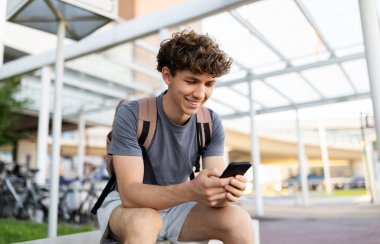 Young Caucasian man with backpack sitting at airport terminal, smiling at smartphone. Modern lifestyle, travel waiting moments, connection and youth culture