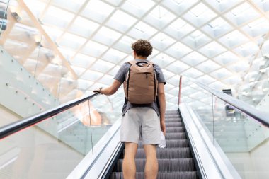 Young Caucasian man with backpack holding water bottle on airport escalator. Represents travel routine, youth journey lifestyle, mobility and passenger waiting