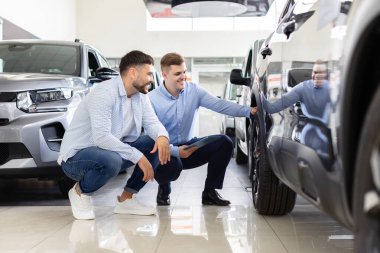 Car salesman and potential customer crouch beside sleek vehicle in modern dealership showroom. Manager holding digital tablet while explaining cars features, man attentively listening
