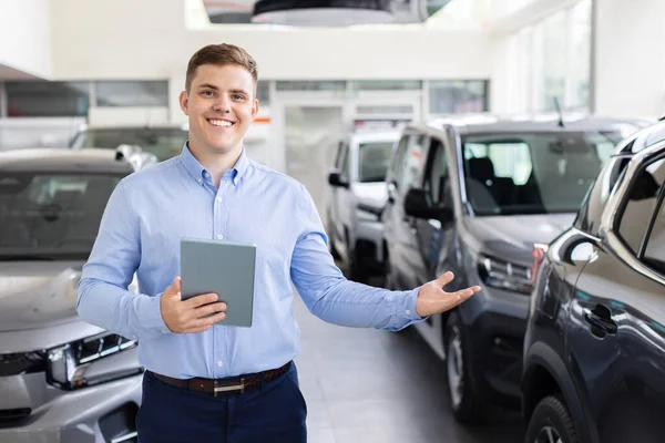 Smiling car dealer presenting car models in showroom. Young professional man holding tablet while standing between new vehicles, lifestyle and transportation theme
