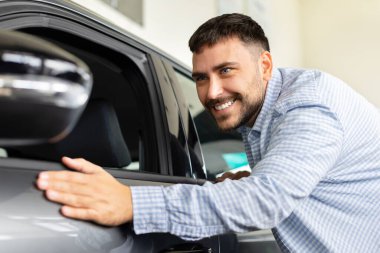 Happy man touching new car at dealership showroom, smiling customer checking vehicle exterior before purchase. Lifestyle, transport and consumer satisfaction