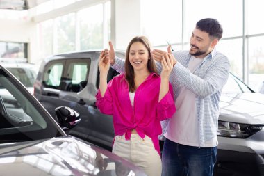 Loving man making surprise to his wife with buying car, covering her eyes. Joyful young couple celebrating their new car purchase in a modern dealership showroom