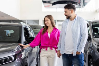 Smiling couple touching new car at dealership showroom, man and woman holding hands while examining vehicle. Lifestyle, consumer choice and automobile purchase concept