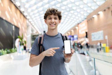 Young Caucasian man with backpack at airport pointing at blank smartphone screen. Concept of digital promotion, travel lifestyle, youth culture and technology