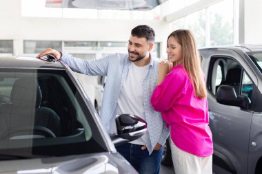 Young caucasian couple smiling while looking at car in dealership showroom, lifestyle moment of auto shopping, vehicle choice and modern transport purchase decision