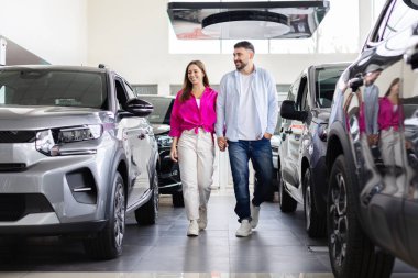 Smiling couple walking inside showroom after car purchase, happy man and woman holding hands between vehicles at dealership. Lifestyle, transport and automobile shopping