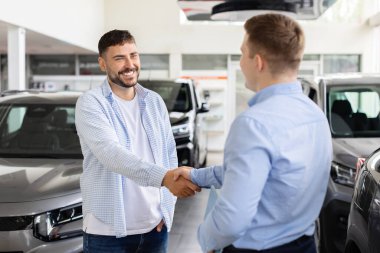 Smiling man shaking hands with car dealer at dealership, customer finalizing automobile purchase near vehicles. Lifestyle, agreement, transport and consumer trust