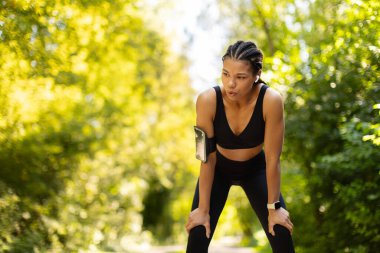 Tired but determined African American woman catching breath after running outdoors. Symbol of strength, resilience, motivation and active lifestyle in park.